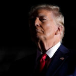 A political figure in a dark setting, wearing a suit and red tie, with a serious expression