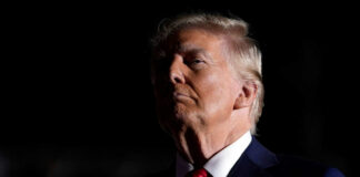 A political figure in a dark setting, wearing a suit and red tie, with a serious expression
