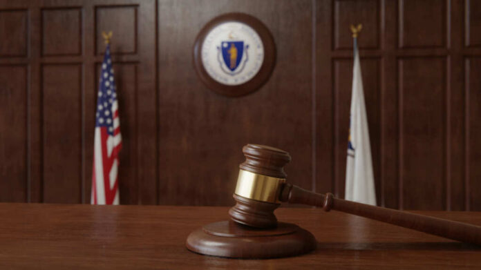 A wooden gavel resting on a table in a courtroom with flags in the background