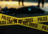 Yellow police caution tape at a crime scene with blurred figures in the background