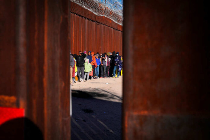 A line of people waiting near a border wall with barbed wire
