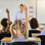 Children in a classroom raising their hands to answer a question while a teacher stands in front