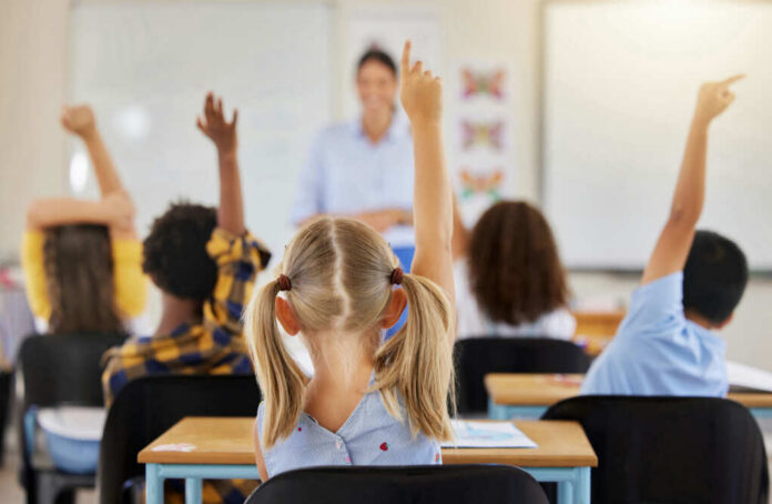 Children in a classroom raising their hands to answer a question while a teacher stands in front