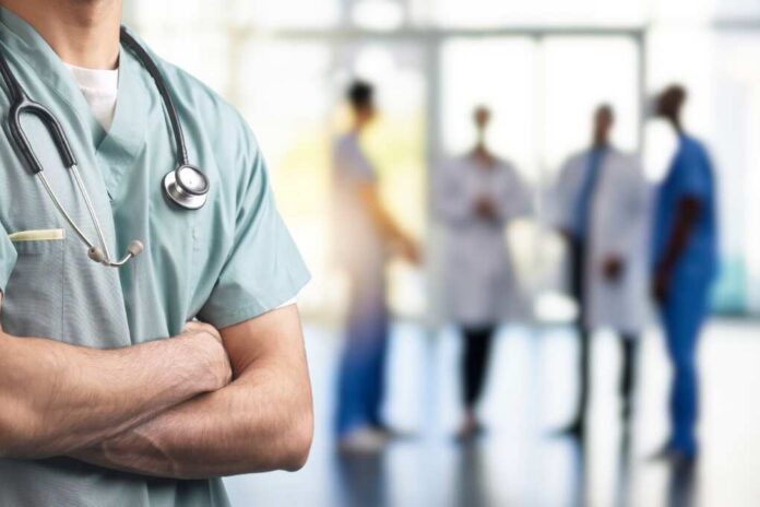 A healthcare professional in scrubs with a stethoscope, standing with arms crossed in a hospital setting