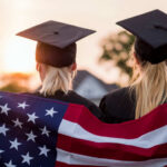 Two graduates wearing caps holding an American flag during sunset