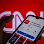 A hand holding a smartphone displaying the CNN news app against a red background
