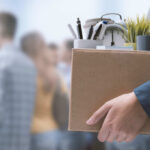 A person holding a cardboard box filled with office supplies and a newspaper