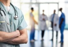 A healthcare professional in scrubs with a stethoscope, standing with arms crossed in a hospital setting