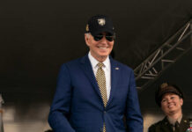 A smiling man in a suit and sunglasses at a military event, with a woman in uniform beside him