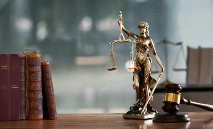 shutterstock2483357455jpg A bronze statue of Lady Justice holding scales and a sword, with law books and a gavel in the foreground