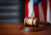 A wooden gavel resting on a round base with a blurred American flag in the background