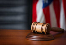 A wooden gavel resting on a round base with a blurred American flag in the background
