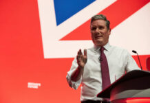 A man in a white shirt and tie gestures while speaking at a podium with a British flag backdrop