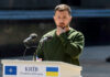 A public figure in a green sweater pondering during a speech at a podium in Kyiv