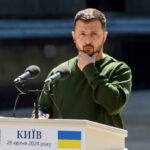 A public figure in a green sweater pondering during a speech at a podium in Kyiv