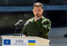 A public figure in a green sweater pondering during a speech at a podium in Kyiv