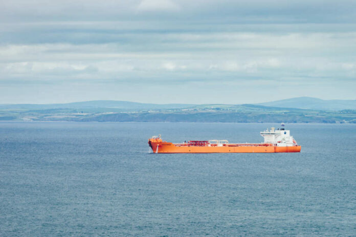 An orange cargo ship sailing in the ocean with a coastal landscape in the background