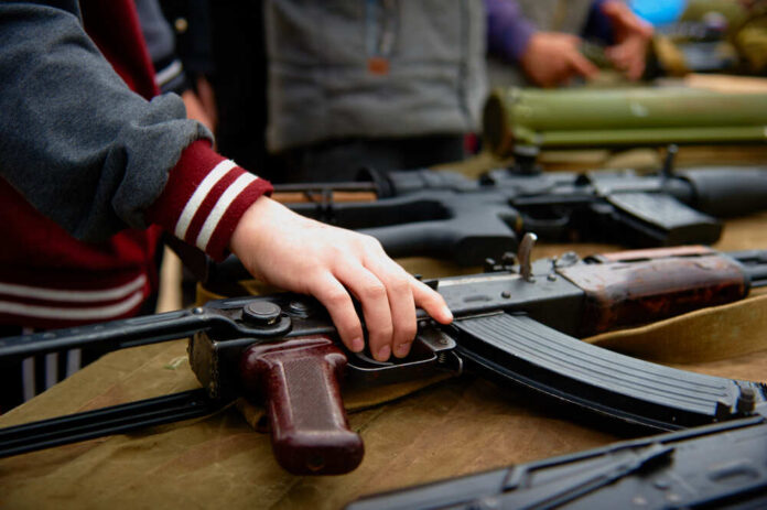 A,Child's,Hand,Takes,An,Automatic,Rifle,From,The,Table Hand touching assault rifle on display table