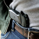 A handgun partially visible in a belt holster worn by a person in casual clothing