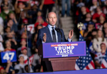 A speaker addressing a crowd at a political rally with campaign signs and flags in the background