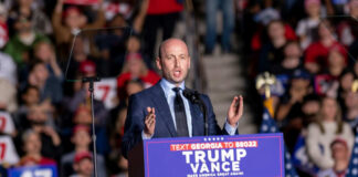 A speaker addressing a crowd at a political rally with campaign signs and flags in the background