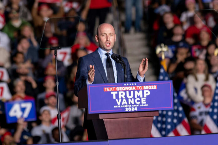 A speaker addressing a crowd at a political rally with campaign signs and flags in the background