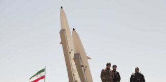 Military personnel standing near missile launchers with an Iranian flag in the background