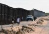 Border fence with patrol vehicles and people walking on a dirt road