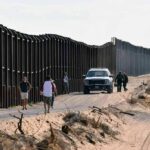 Border fence with patrol vehicles and people walking on a dirt road