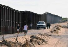Judge Greenlights Wall Through Tribal Lands Border fence with patrol vehicles and people walking on a dirt road