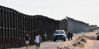 Border fence with patrol vehicles and people walking on a dirt road