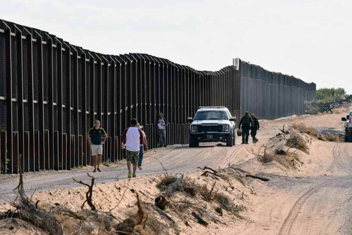 Border fence with patrol vehicles and people walking on a dirt road