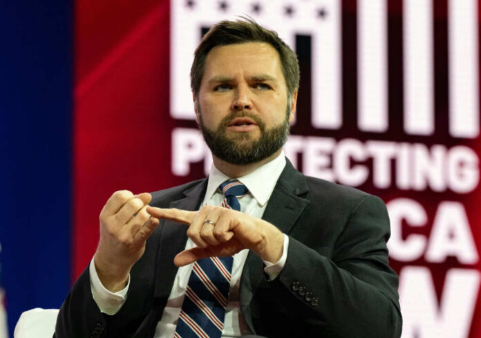 Man in suit speaking on stage with red and white backdrop