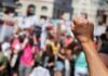 Tear Gas Floods Austin After ICE Gunfire Raised fist in foreground at a crowded street protest