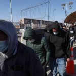 People in hoodies and masks walk in a line along a fenced walkway