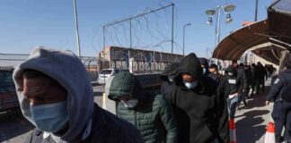 People in hoodies and masks walk in a line along a fenced walkway