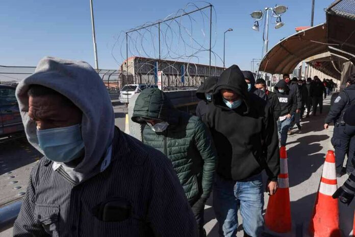 People in hoodies and masks walk in a line along a fenced walkway