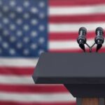 Podium with microphones in front of a blurred American flag