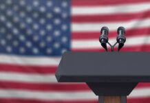 Podium with microphones in front of a blurred American flag