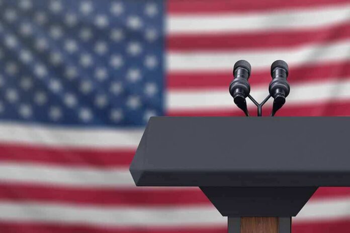 shutterstock_1223942518 Podium with microphones in front of a blurred American flag