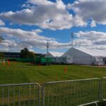 Temporary white tents and equipment on a grassy field with traffic cones and metal barriers